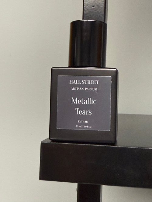 Perfume bottle labeled  Metallic Tears by Hall Street Artisan Parfum on a dark surface with a neutral background.